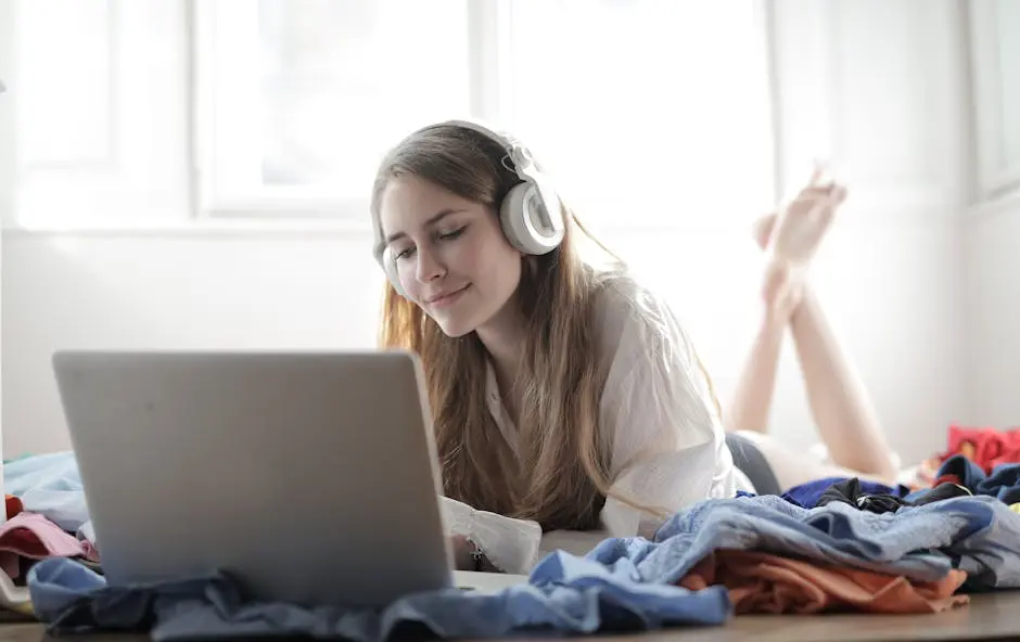 Young woman enjoying music and working on laptop in a cozy bedroom setting.